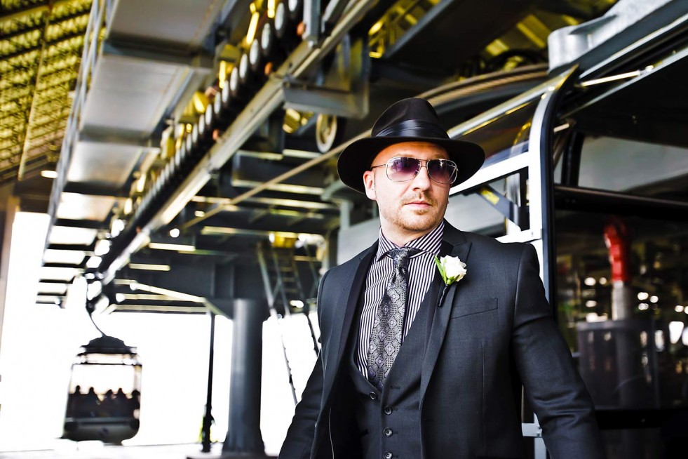 A man in a black suit, striped shirt, and black hat with sunglasses is standing near a cable car station. He has a white flower on his lapel. In the background, a cable car can be seen, and the station structure is visible—capturing moments like this could elevate any Colorado wedding photography session.