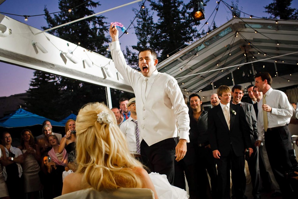 A man in a white shirt enthusiastically raises an item in the air, while a blonde woman in a white dress sits in the foreground. Several people in formal attire stand around them, smiling and cheering, under a canopy decorated with string lights—a perfect capture for destination wedding photography.