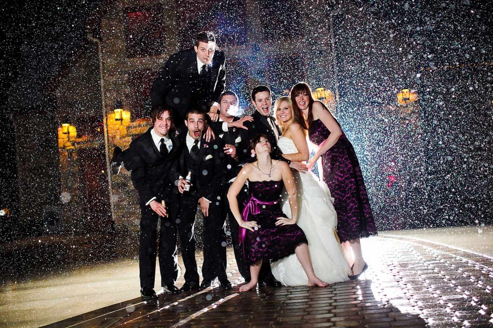 A wedding party poses for a photo under falling snow, showcasing the charm of Colorado wedding photography. They wear formal attire, with the bride in a white gown and groom in a black suit. Four bridesmaids in purple dresses and three groomsmen in black suits surround the smiling couple.