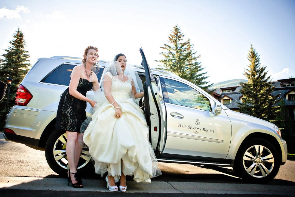 A bride in a white wedding dress is stepping out of a silver SUV with the help of another woman in a black dress. The SUV has "Four Seasons Resort" written on its side. Trees and a building are visible in the background under a clear, blue sky—perfect for Colorado wedding photography.