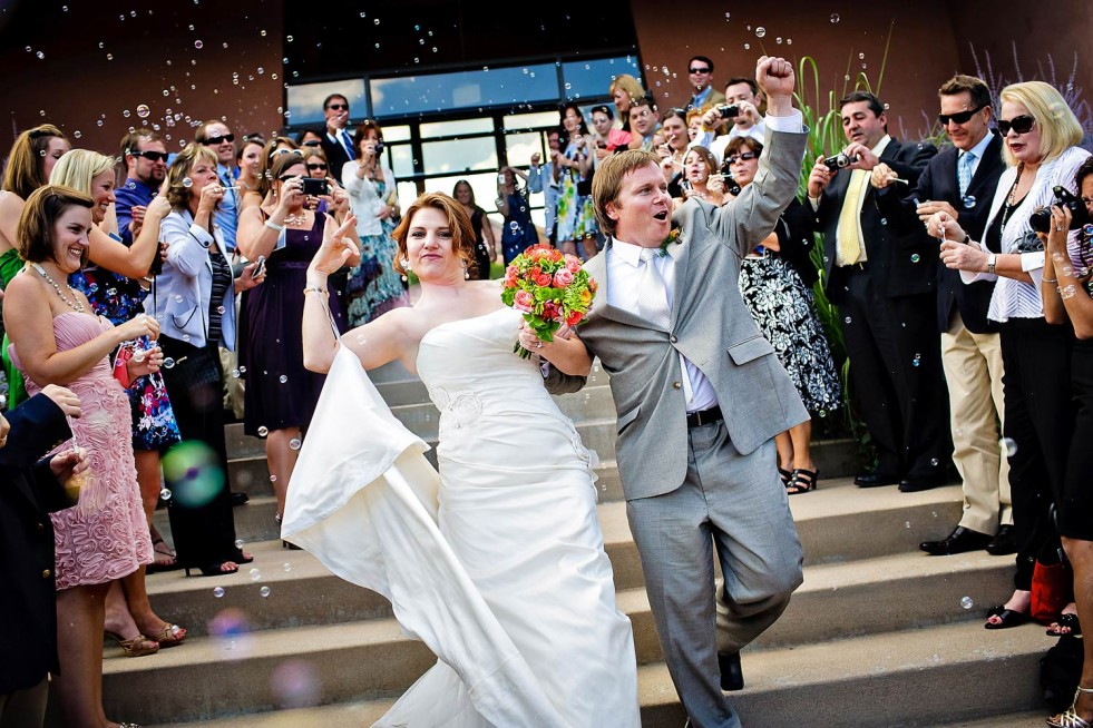 A newlywed couple walks down the stairs outside their wedding venue, surrounded by cheering guests blowing bubbles. The bride is in a white gown holding a bouquet, while the groom is in a gray suit, both with enthusiastic expressions. This perfect moment captures the essence of Colorado wedding photography.